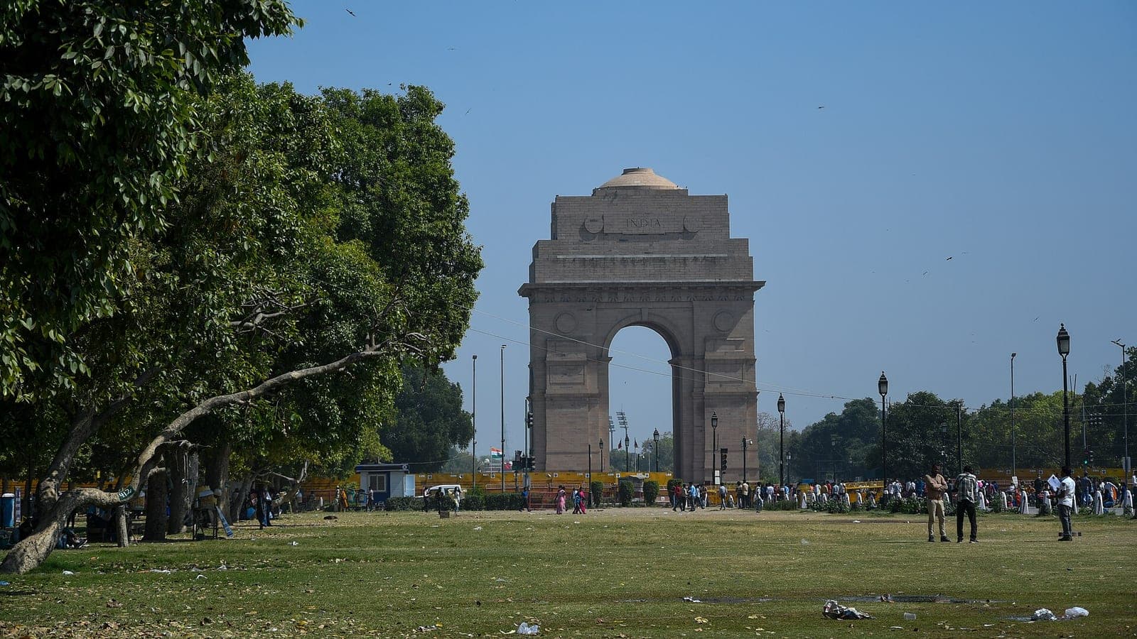 India Gate viewed across the lawns of Kartavya Path, framed by a spreading tree, with people gathered at its base.