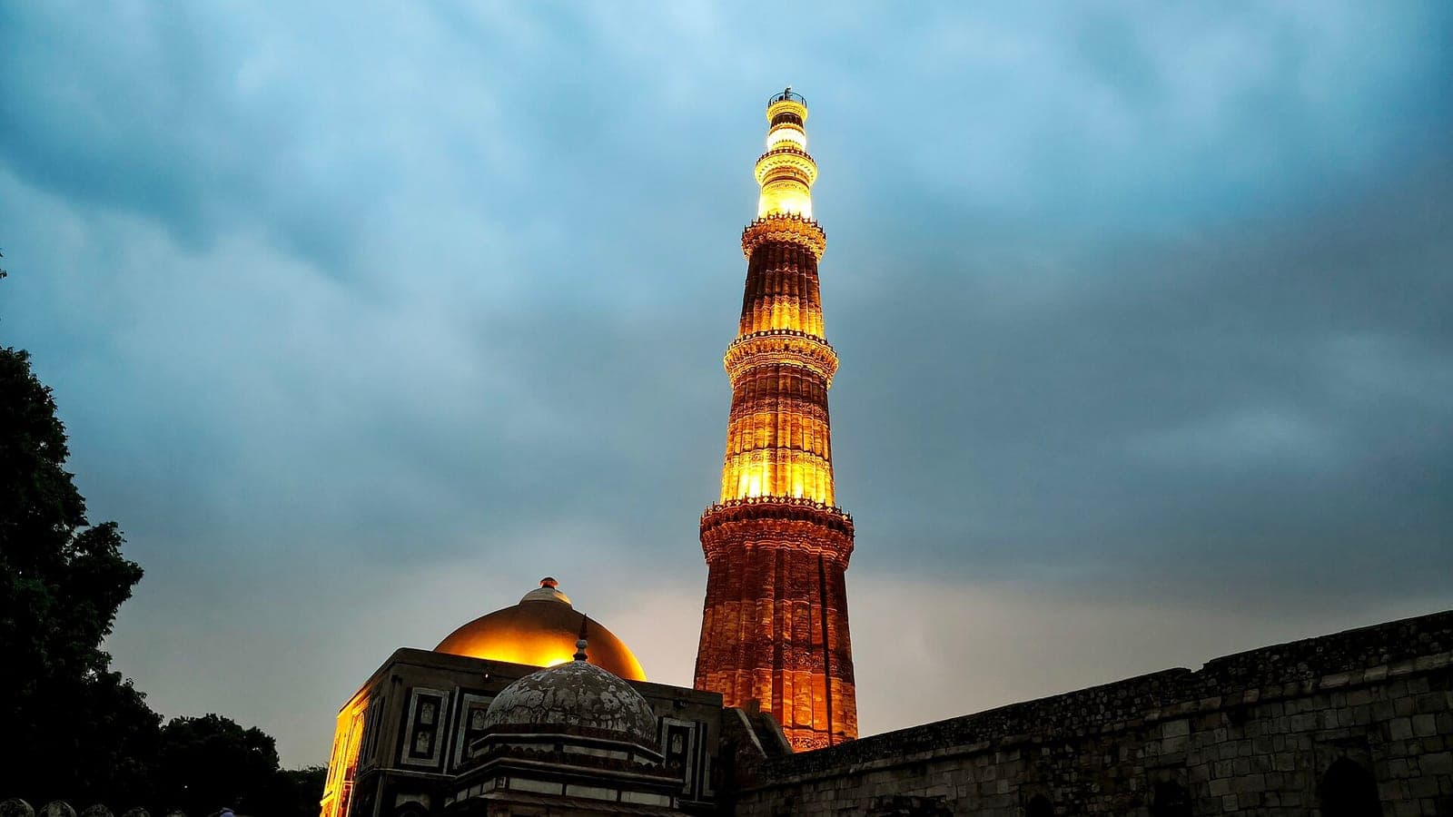 The Qutub Minar lit at dusk, its fluted sandstone storeys tapering against a stormy sky.